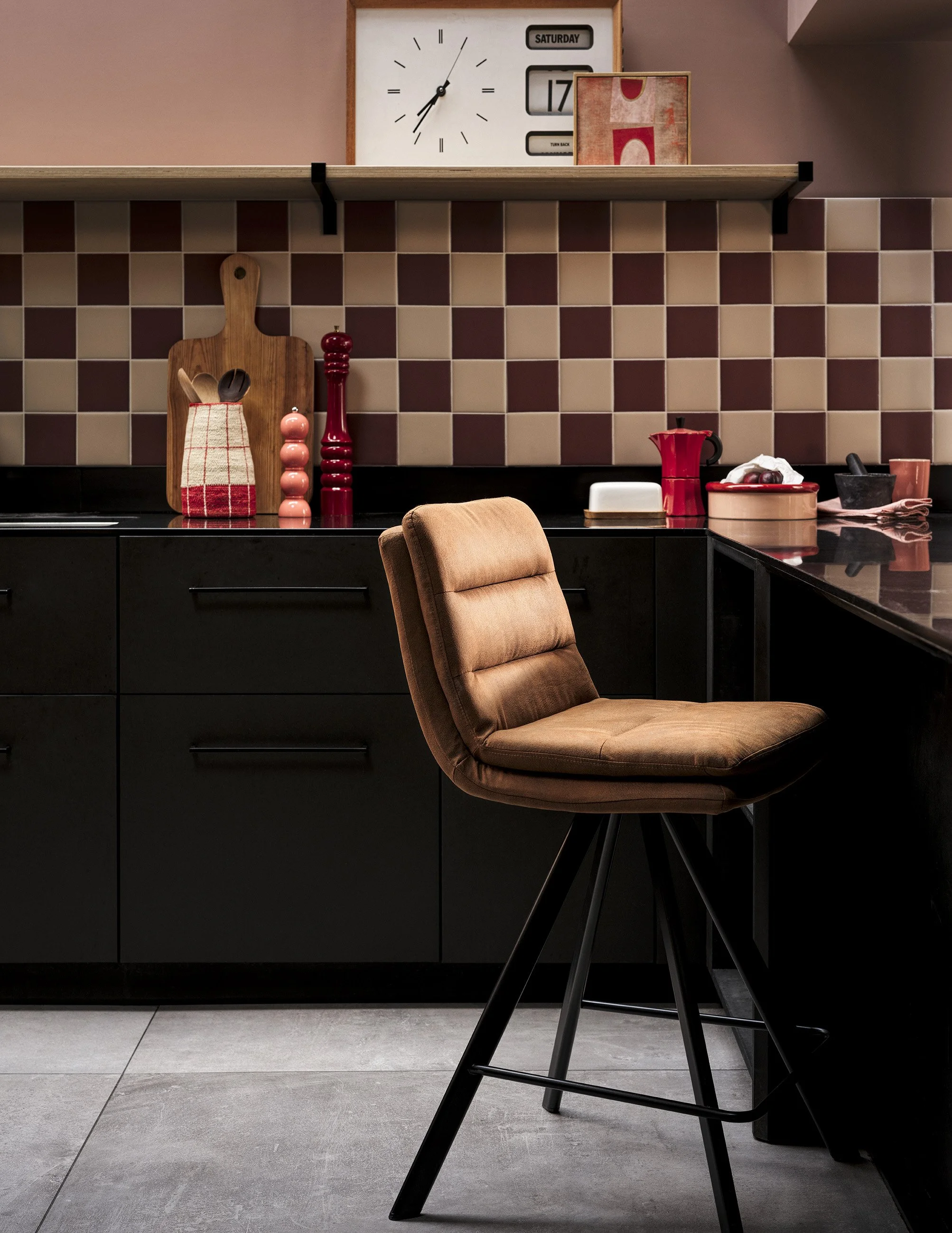 A kitchen scene featuring a beige swivel chair in the foreground, black cabinets, a tiled red and beige backsplash, and a wooden shelf with a clock, calendar, and decorative items.