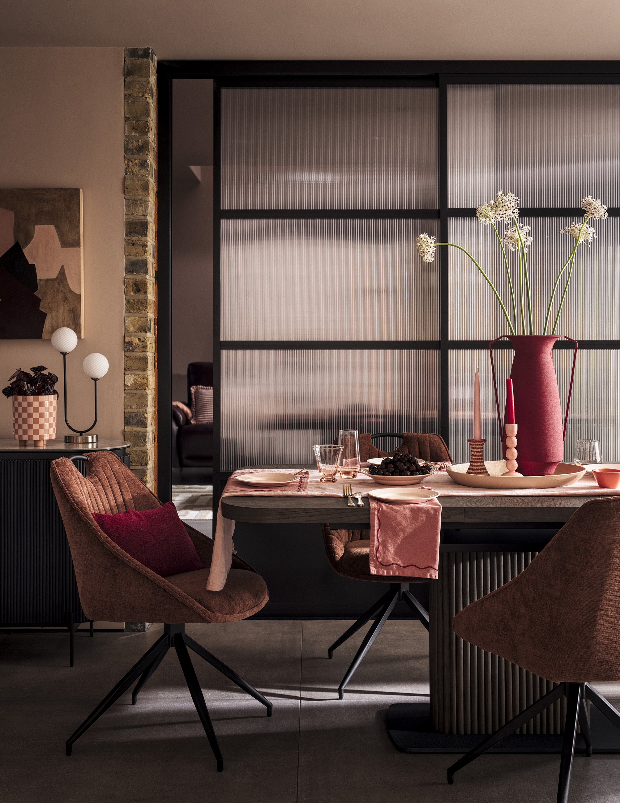A dining room with a large pink vase filled with white flowers on the table, surrounded by pink and brown chairs, and decorated with pink and red accents.
