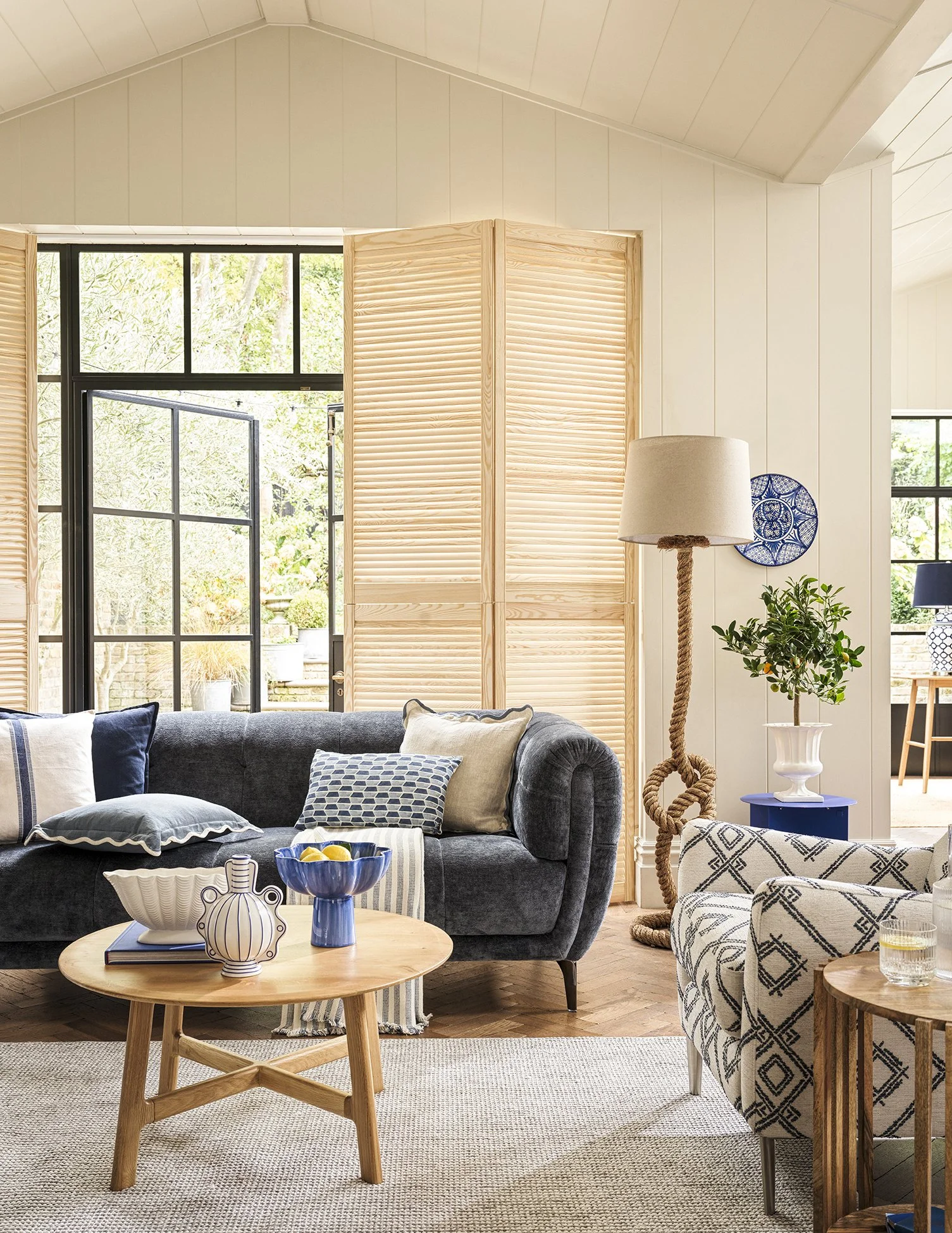 Living room with dark gray sofa, wooden coffee table with vases, armchair with patterned fabric, window with view of greenery, light wood folding screen, floor lamp with rope detail, and decorative items.