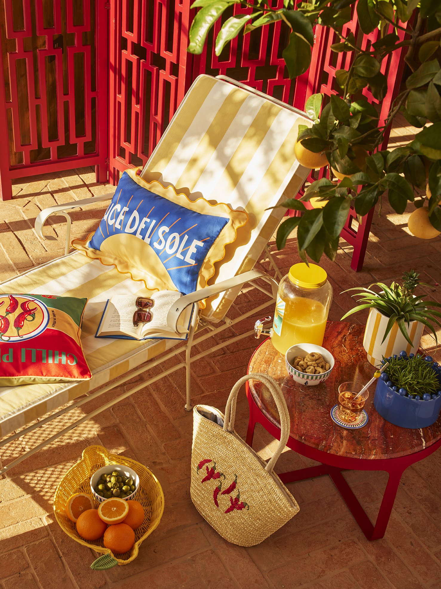 Colorful outdoor scene with a lounge chair, pillows, an open book, sunglasses, a small table with a pitcher of orange juice, a bowl of cookies, a bowl of green olives, a glass of juice, potted plants, a fruit basket with oranges and green olives, a t