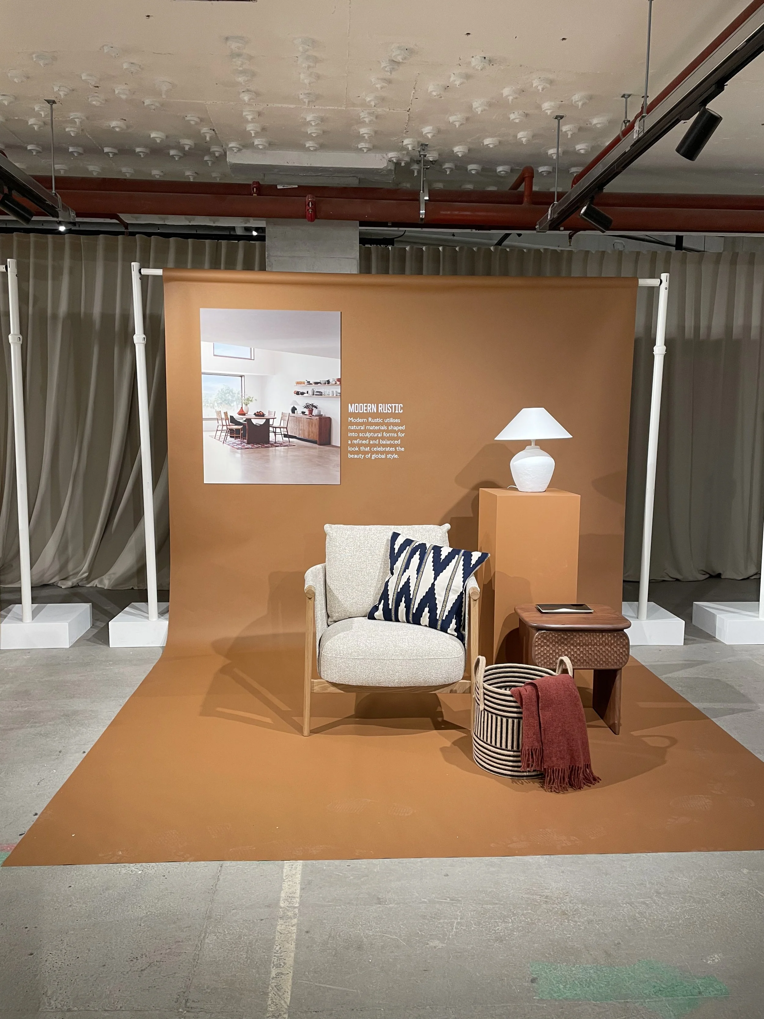 Display of modern rustic living room setup on a brown backdrop, featuring a beige armchair with a navy and white patterned pillow, a side table with a tablet, a table lamp, woven basket, and a red throw blanket, with a sign explaining the style.