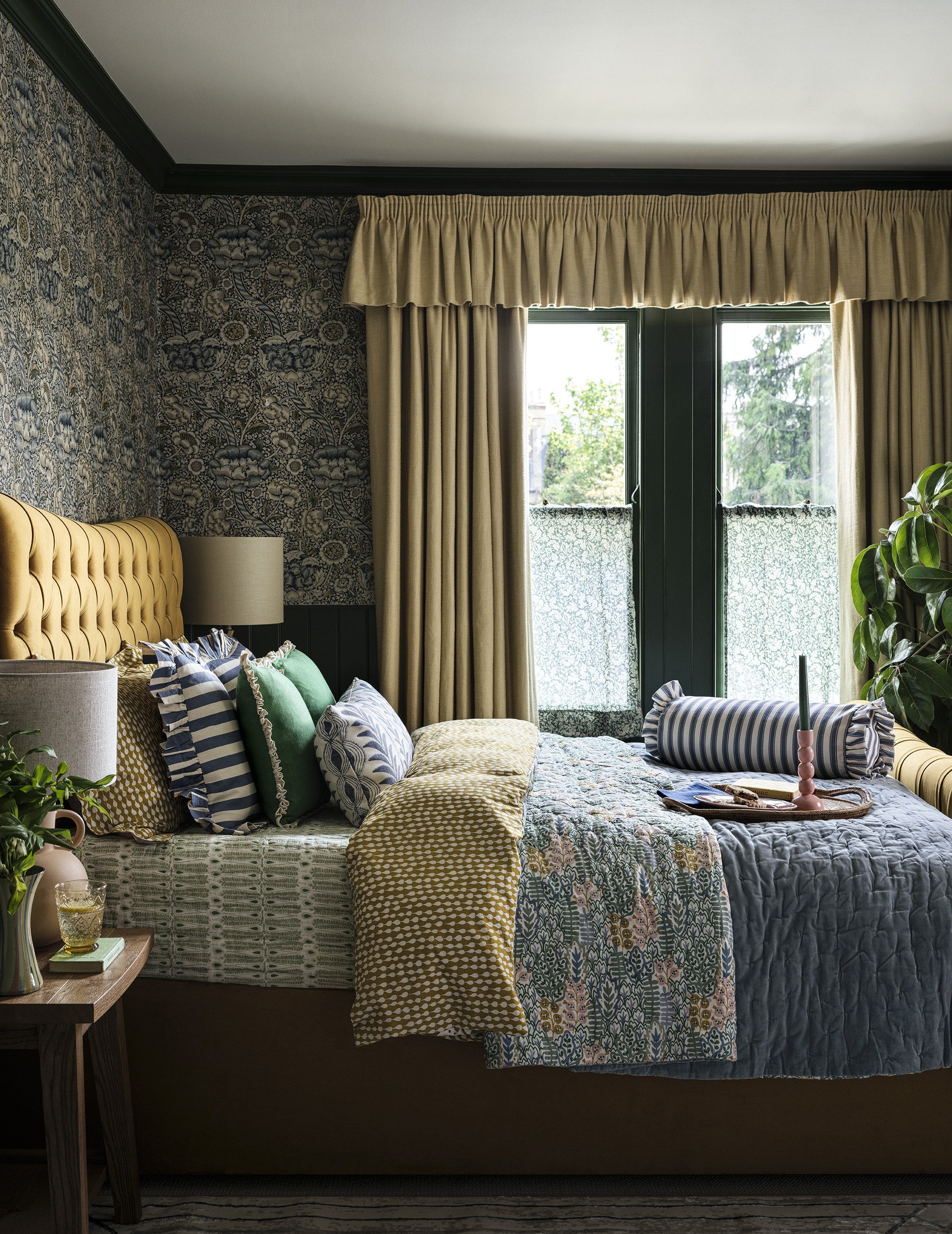 A bedroom with a large bed covered in a multicolored patchwork quilt and decorated with an array of striped and solid pillows in green, blue, and yellow. The headboard is tufted and upholstered in a mustard yellow fabric. Beside the bed, there is a w