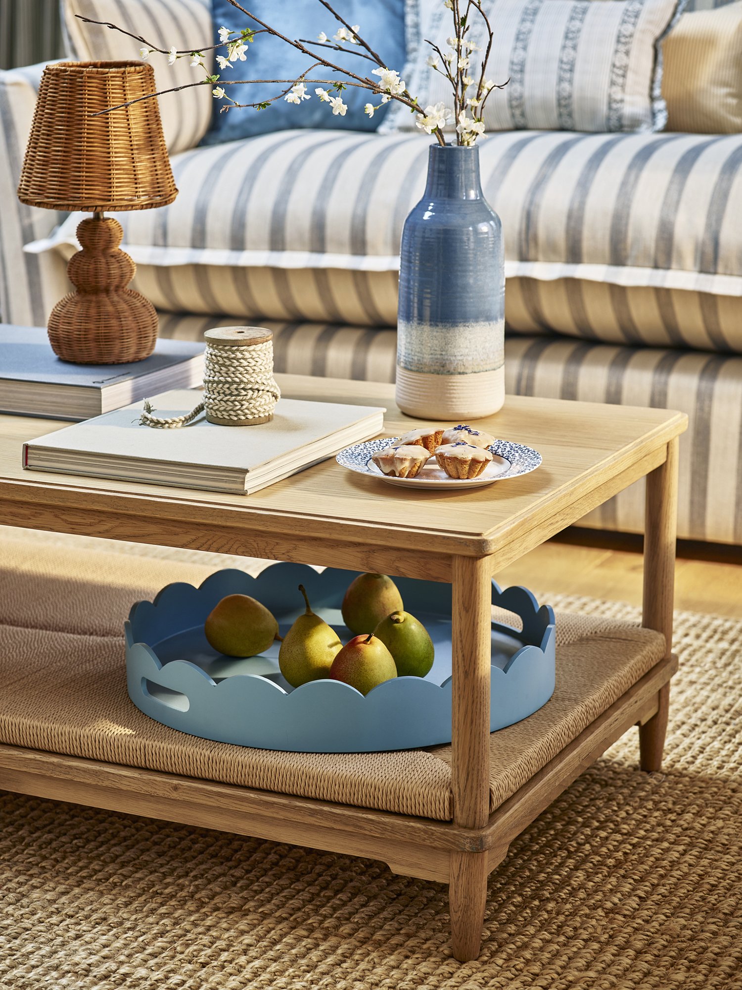A cozy living room with a wooden coffee table, a woven placemat, and a sofa with striped cushions. The table has a blue tray with pears underneath, a tall blue and white vase with cherry blossom branches, a wicker lamp, some books, a spool of rope, a