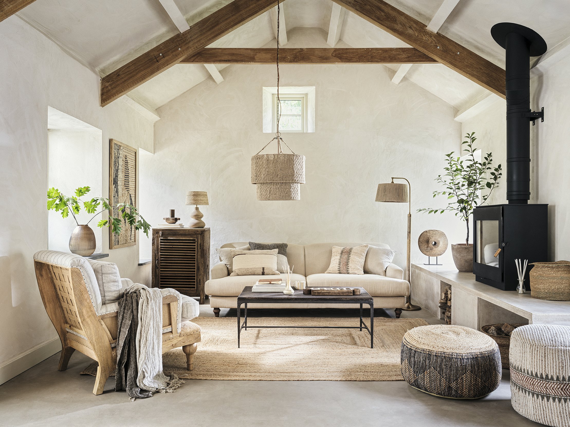 A cozy living room with a vaulted ceiling supported by wooden beams, a white sofa with cushions, a wooden armchair, a black fireplace with a chimney, potted plants, woven ottomans, lamps, and a coffee table with books.