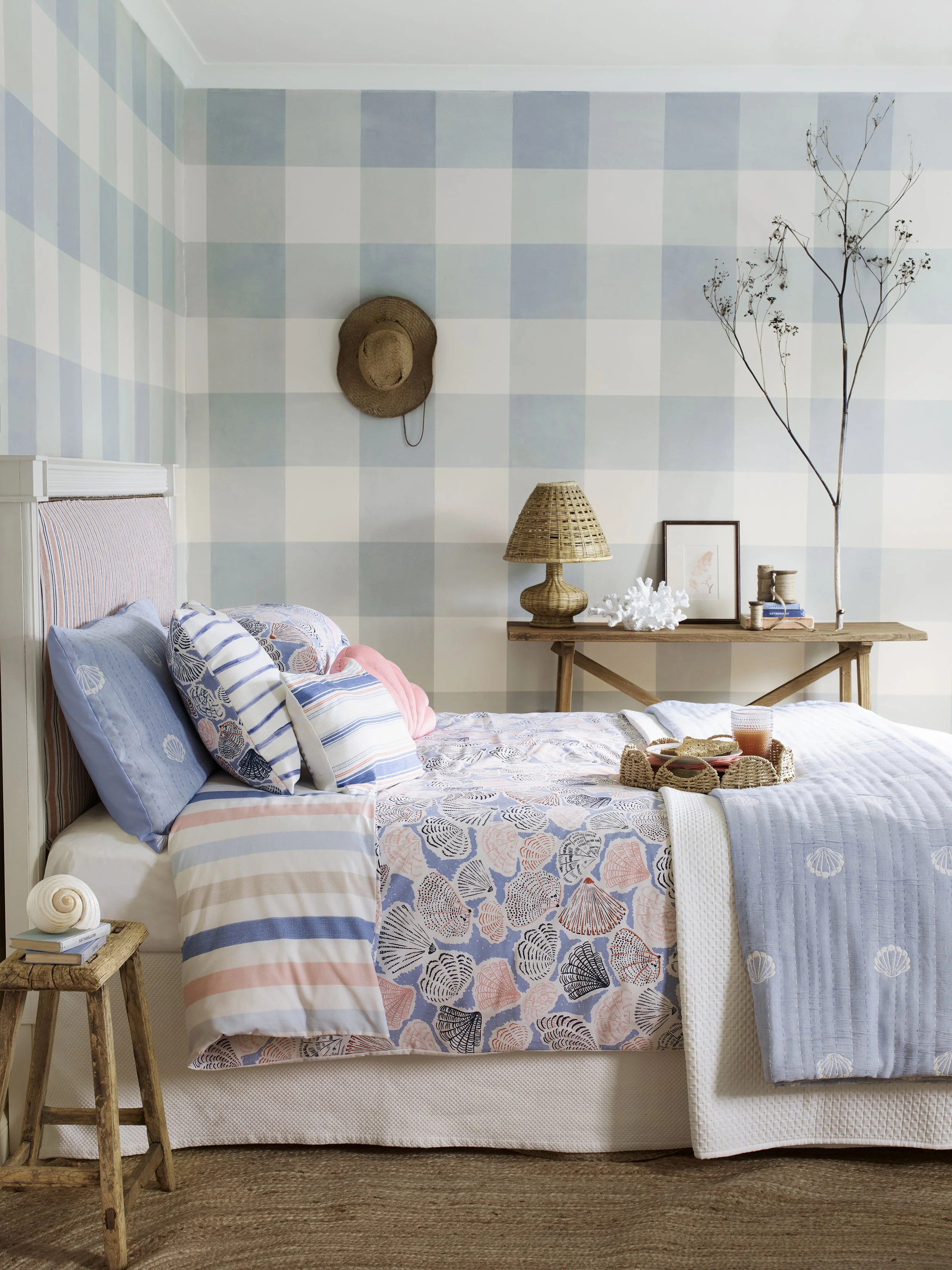 A bedroom with a checkered white and light blue wall, a white bed with beach-themed pillows, a wooden side table with a small stack of books and a shell, and a wooden console table with a wicker lamp, framed art, branches, and coral decor.