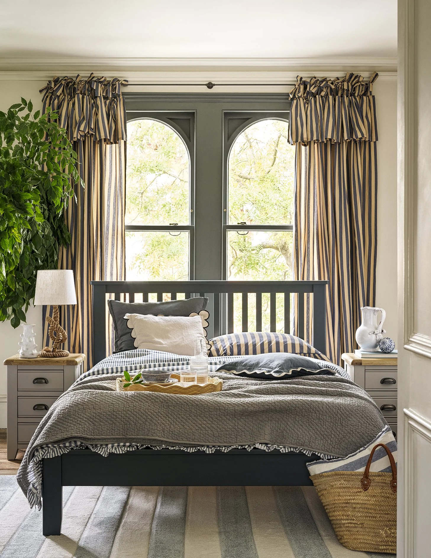 A bedroom with two arched windows, cream walls, a navy bed frame, striped curtains, a beige quilt, and assorted pillows. There are two nightstands with decor, a wicker basket by the bed, and a large green plant to the left.