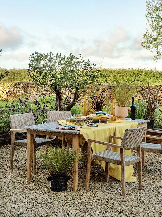 Outdoor table set with yellow tablecloth, surrounded by four chairs and decorated with potted plants and tableware, in a garden or backyard setting.