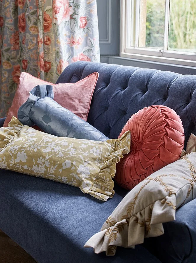 Various decorative pillows arranged on a dark blue upholstered sofa by a window with floral curtains.