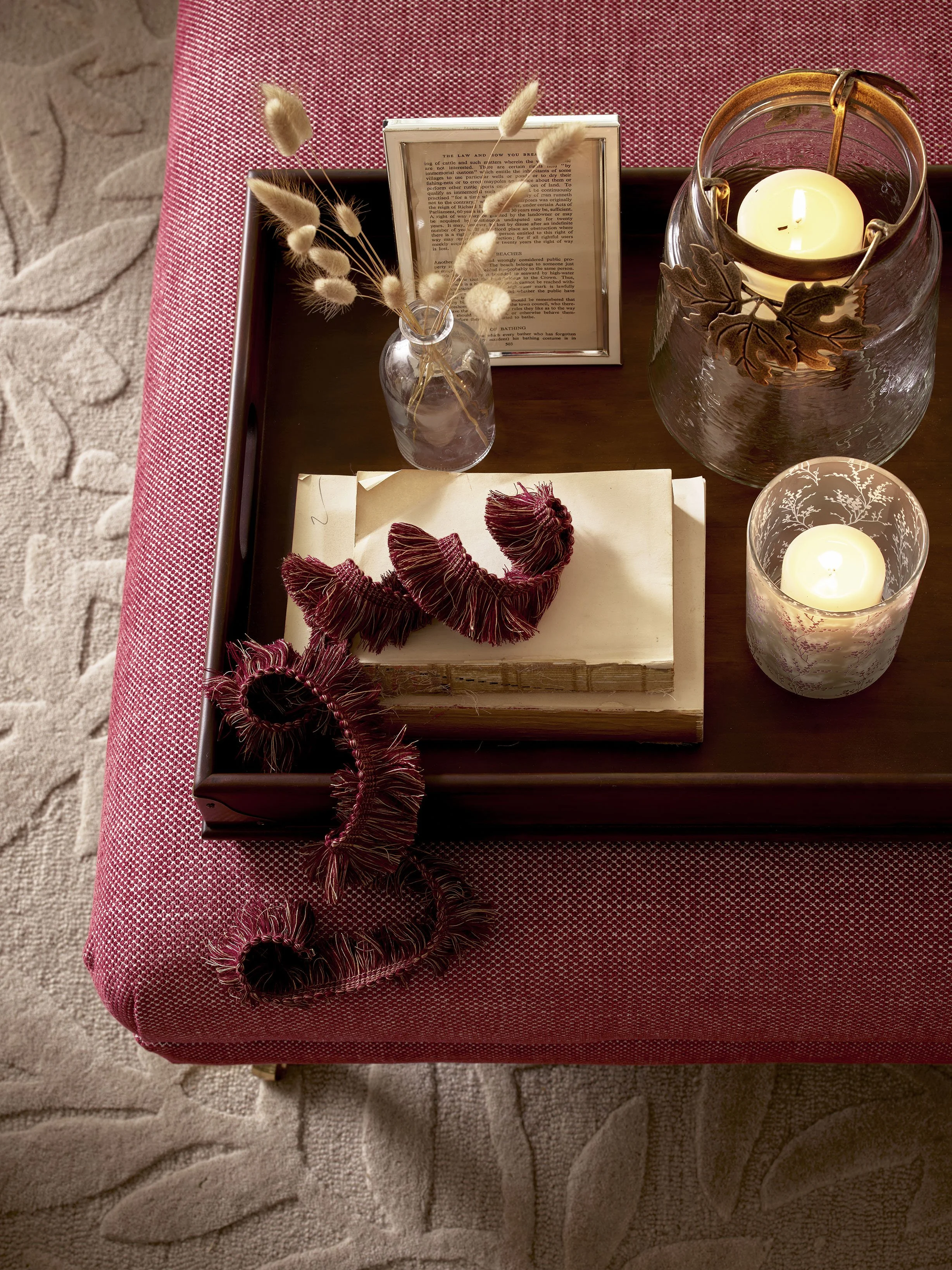 Decorative table setup with candles, a framed photo, and dried flowers on a tray atop a red-patterned upholstered ottoman.