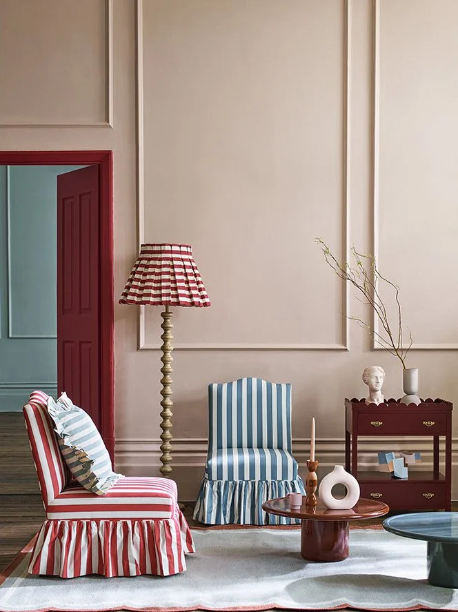 A living room with striped furniture and decor, including a red and white striped armchair with a pillow, a blue and white striped armchair, a side table with a candle and a decorative vase, and a red cabinet with a sculpture and a tall plant. A red 