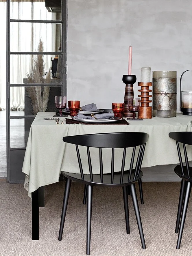 A dining table set with a cream-colored tablecloth, four red glasses, dark grey plates, and silverware. Decorative candles and vases in black, pink, and neutral tones are arranged on the table. Black chairs are positioned around the table, with a sli