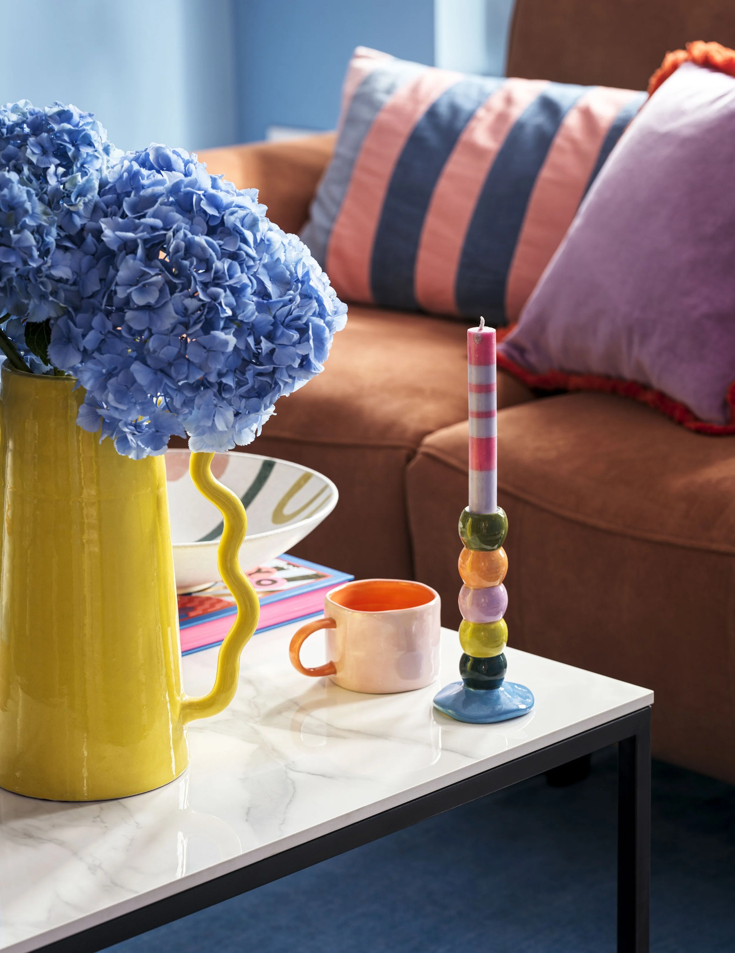 Colorful living room with a blue hydrangea bouquet in a yellow pitcher, a pink and striped candle, a pink mug, and a bowl on a white marble table in front of a sofa with striped and pink pillows.