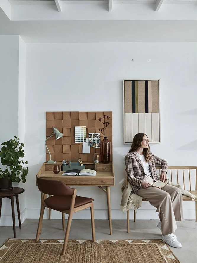 A woman sitting on a wooden bench in a neatly decorated room, holding a book, with a desk and chair nearby, a potted plant, and abstract wall art.