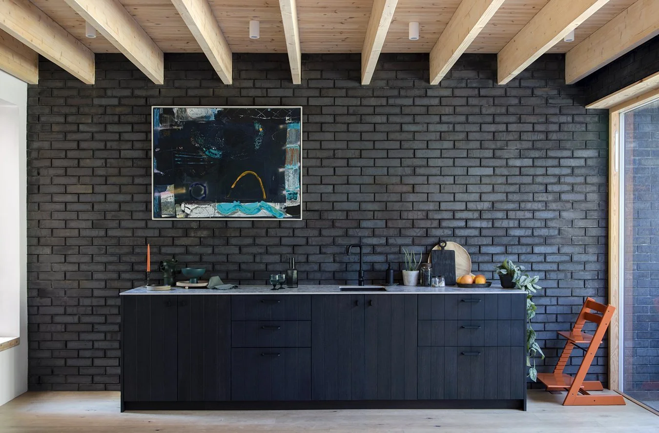 Modern kitchen with black brick wall, wooden ceiling beams, black cabinetry, minimal decor, artwork, plants, and a wooden chair, illuminated by natural light from large window.