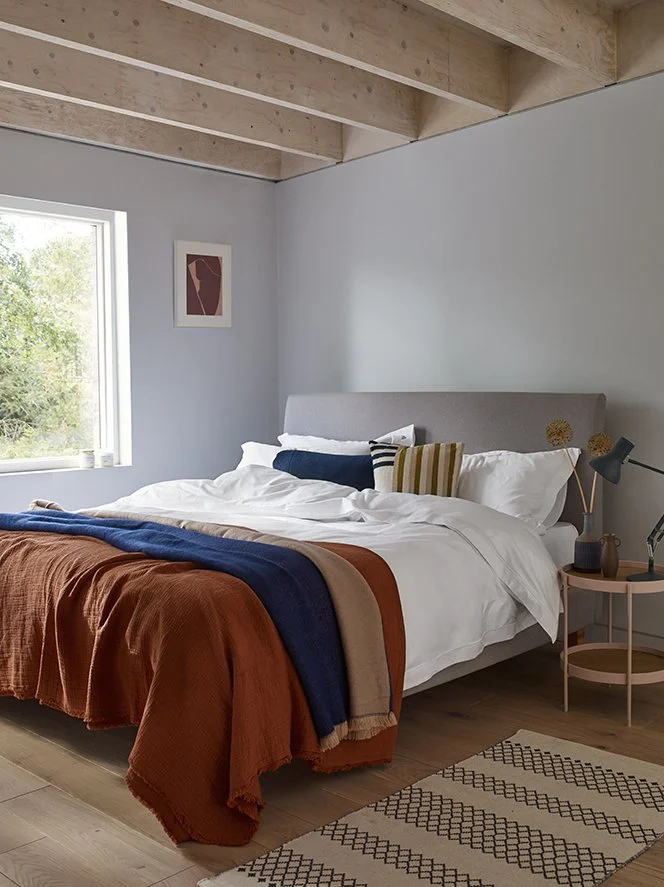 A cozy bedroom with a bed that has white sheets and multiple pillows, including a striped pillow. The bed has a rust-colored blanket with a darker blue blanket on top. Next to the bed is a pink nightstand with a gray desk lamp, a small vase with drie