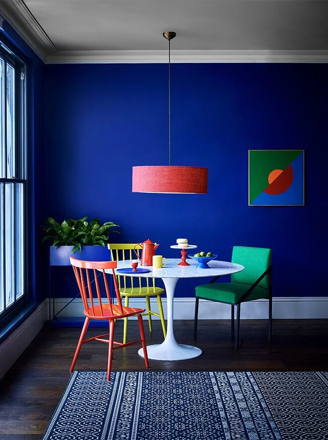 A colorful dining room with a blue wall, a white oval table, and four chairs in red, yellow, green, and turquoise. A large pink drum shade pendant light hangs above the table. A window on the left and a framed abstract art piece on the wall. A green 