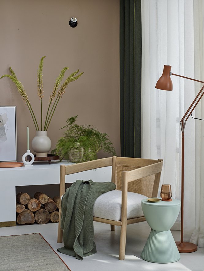 A cozy indoor scene with a light wooden chair, green blanket draped over it, a side table with a green base and a cup, a modern copper floor lamp, a white console table with a white vase holding tall green plants, and a large rock climbing wall on th
