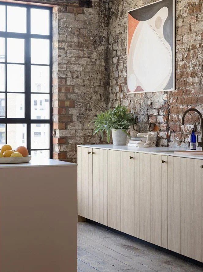Interior of a modern kitchen with exposed brick wall, large window, white cabinetry, a potted plant, a bowl of fruit, and abstract artwork.