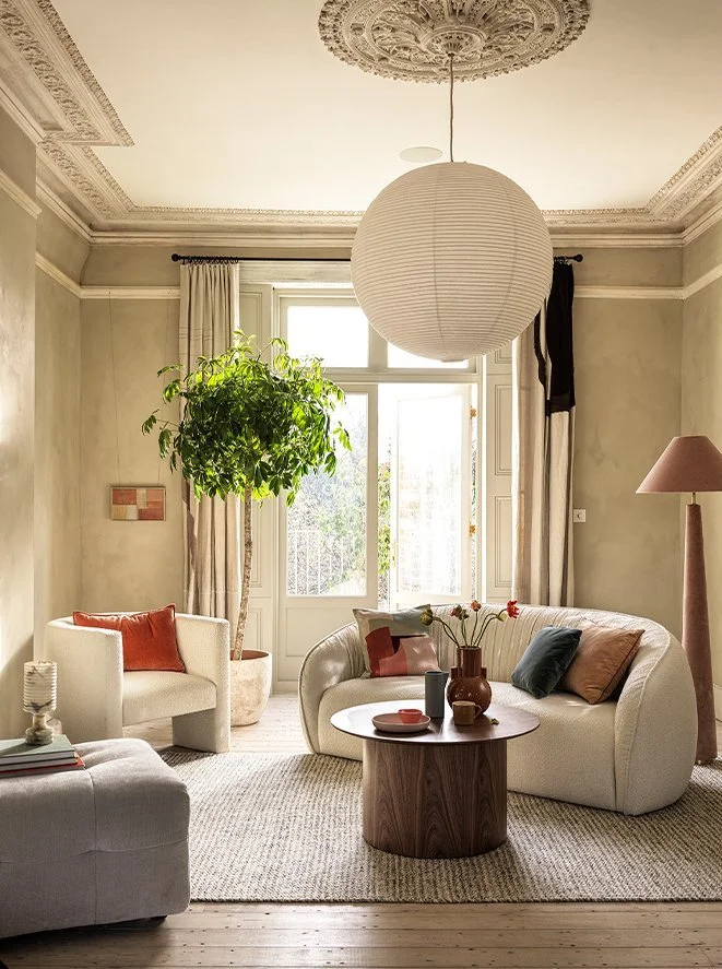 A cozy living room with a large window, cream-colored walls, and elegant ceiling molding. It features a round sofa, a side armchair, a tall floor lamp, and a potted plant hanging from the ceiling. There's a round wooden coffee table with decorative v