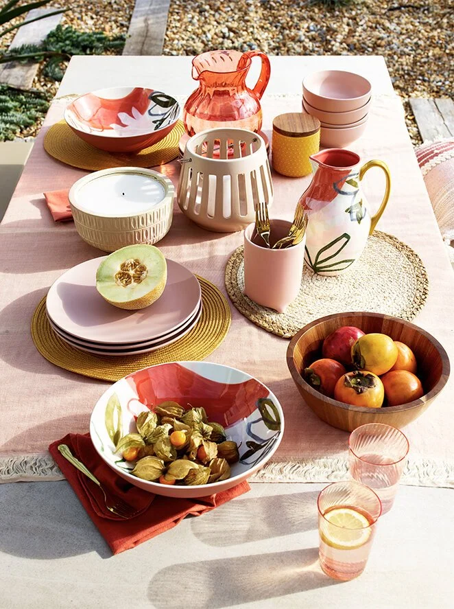 A table set outdoors with pink and white dishes, bowls of fruit, candles, a pitcher, and glasses with lemon slices, on a pink tablecloth with a yellow placemat and other colorful accents.