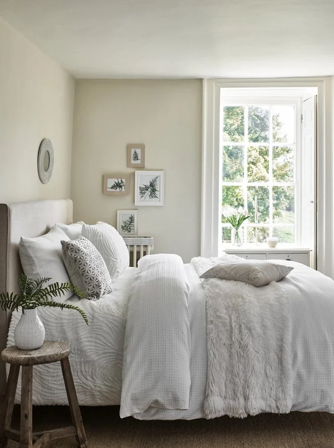 Bright bedroom with white bedding, a small wooden stool with a vase of green ferns, framed botanical art on the wall, and a large window with a vase and small plant on the windowsill.