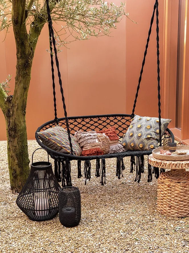 A black outdoor swing with pillows hanging from a black rope, located next to a tree. Below the swing are two black lanterns, one large and one small. To the right, there is a woven round side table with candles and decorative items, set on a gravel 