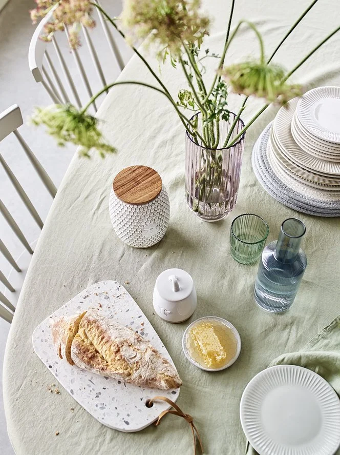 A table set with a green tablecloth, featuring a sliced bread or pastry on a white speckled cutting board, a small dish of honey or jam, a white ceramic container, a tall pink glass vase with fresh flowers, clear and green glass cups, a tall glass bo