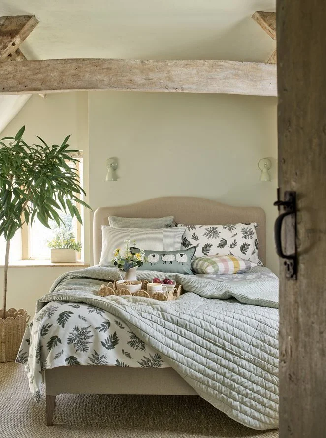 A cozy bedroom with a bed covered in botanical-patterned bedding, a small tray with breakfast foods, a large leafy plant near a window, and a rustic wooden beam on the ceiling.