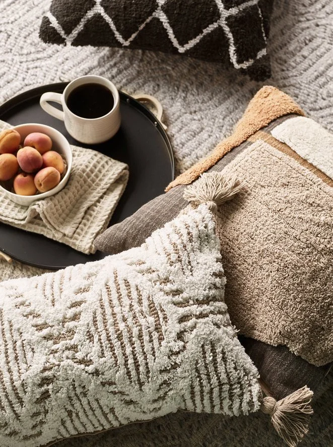 A cozy scene featuring a tray with a cup of coffee, a bowl of peaches, and a cloth napkin on a textured blanket, surrounded by plush pillows and blankets.