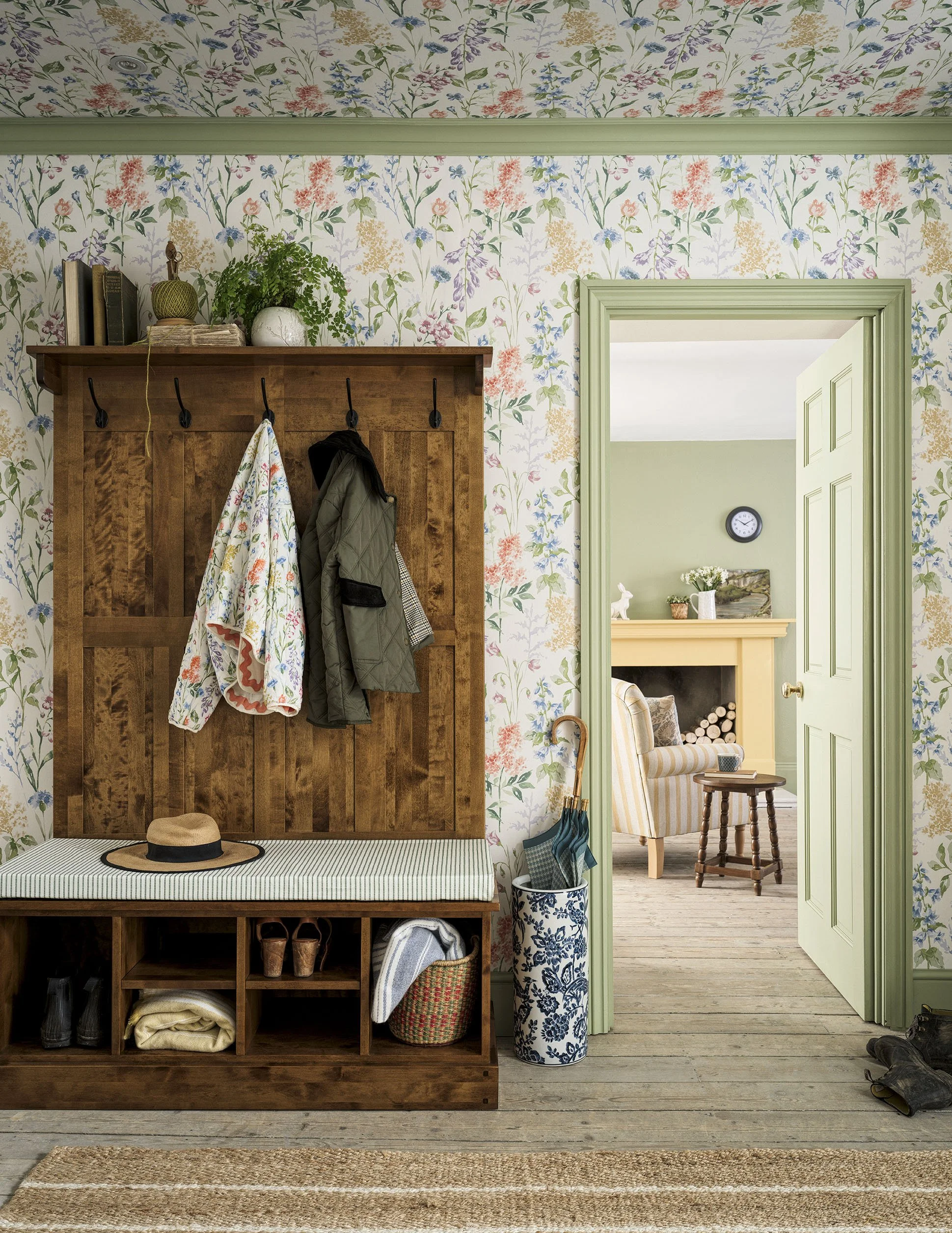 A cozy entryway with a wooden coat rack holding a floral blanket and a green jacket, a cushioned bench with a hat on top, and shoes stored in cubbies. An umbrella stand with umbrellas is near an open doorway leading to a living room with chairs, a fi