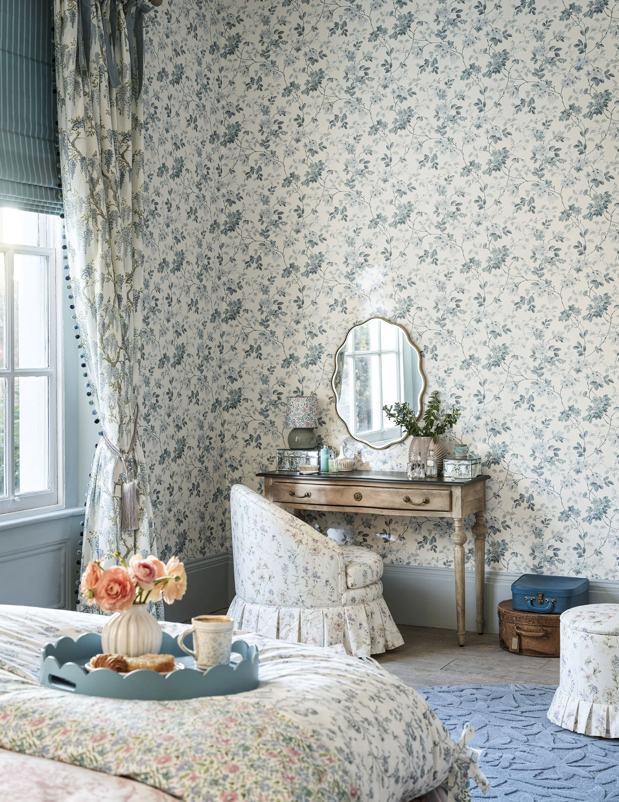 A bedroom with floral wallpaper, a window with curtains, a vintage vanity table with a mirror, a chair, a vase of flowers, and a tray with food and drink on the bed.
