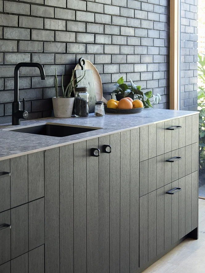 Modern kitchen with gray brick wall, gray countertop, and wooden cabinets. Items on the countertop include potted plants, jars, a cutting board, and a bowl of oranges and lemons.
