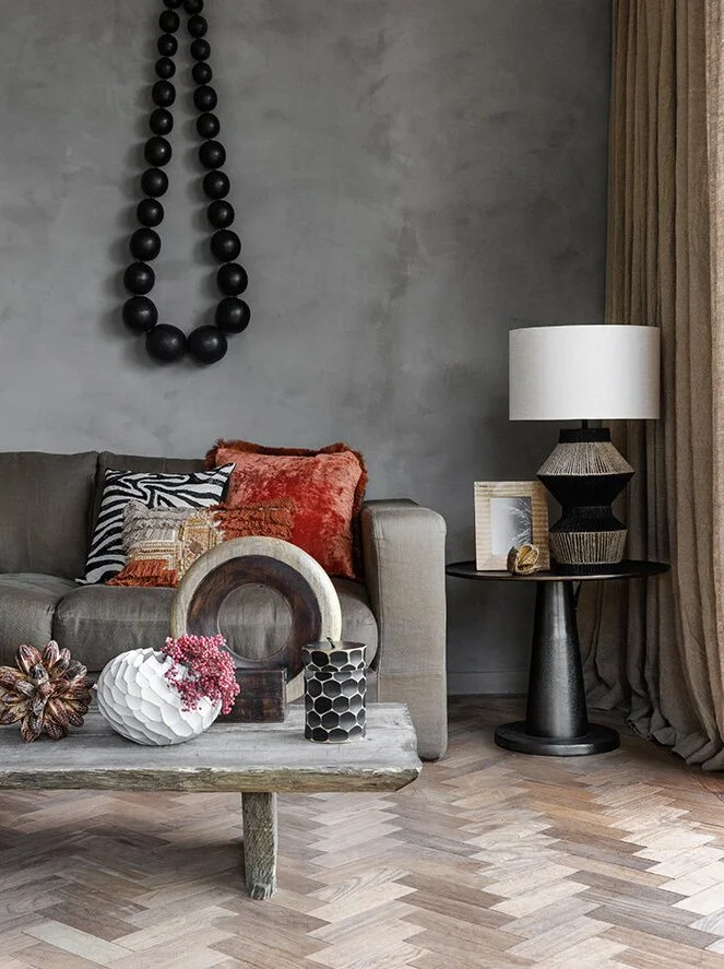 Living room with a gray sofa decorated with patterned and orange pillows, a rustic coffee table with decorative vases, a black side table with a lamp, framed picture, and a floral sculpture, gray textured wall with black bead wall art, and beige curt