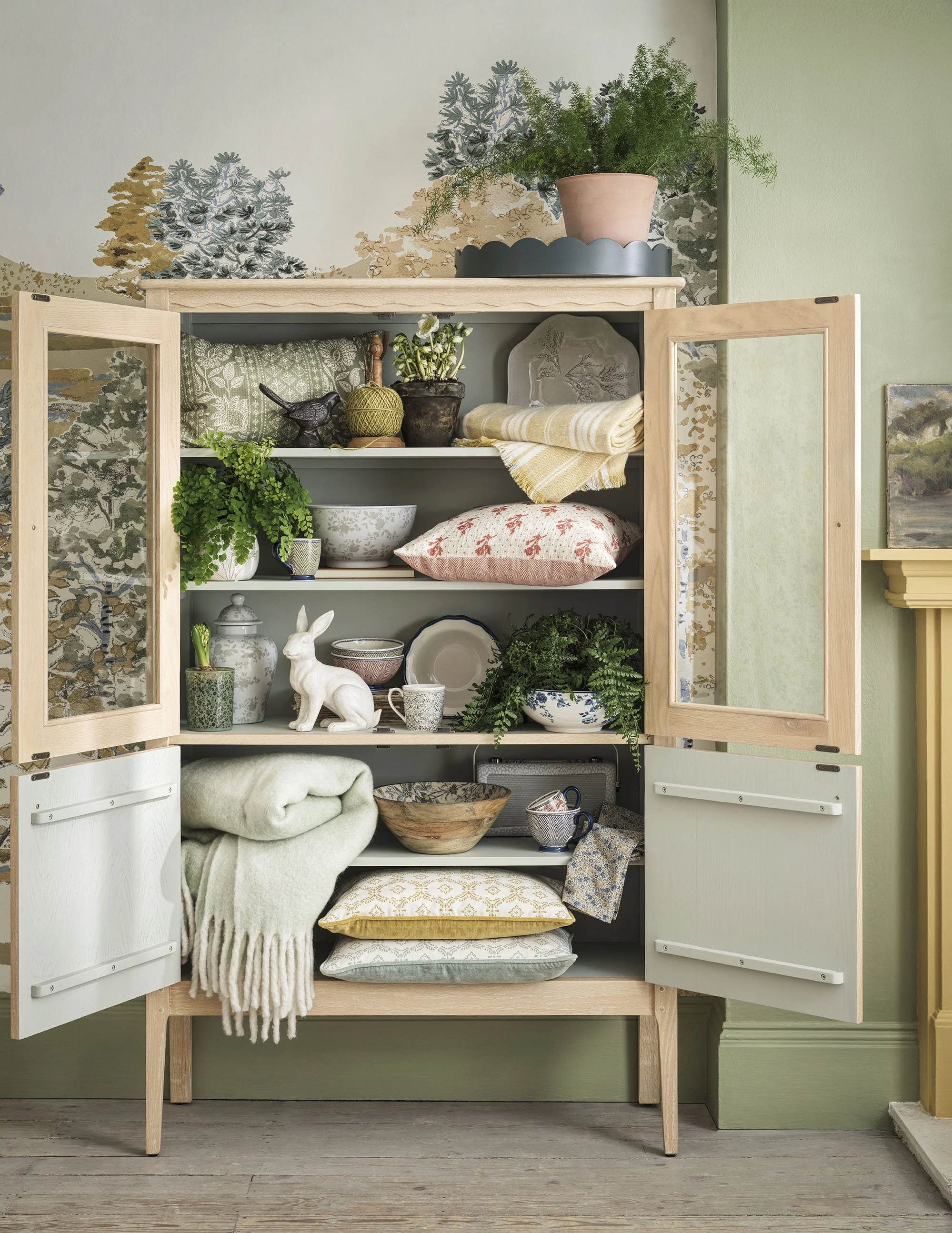 Open wooden cabinet filled with decorative ceramics, bowls, plants, cushions, throws, and a rabbit figurine, with a green wall and nature-themed wallpaper in the background.