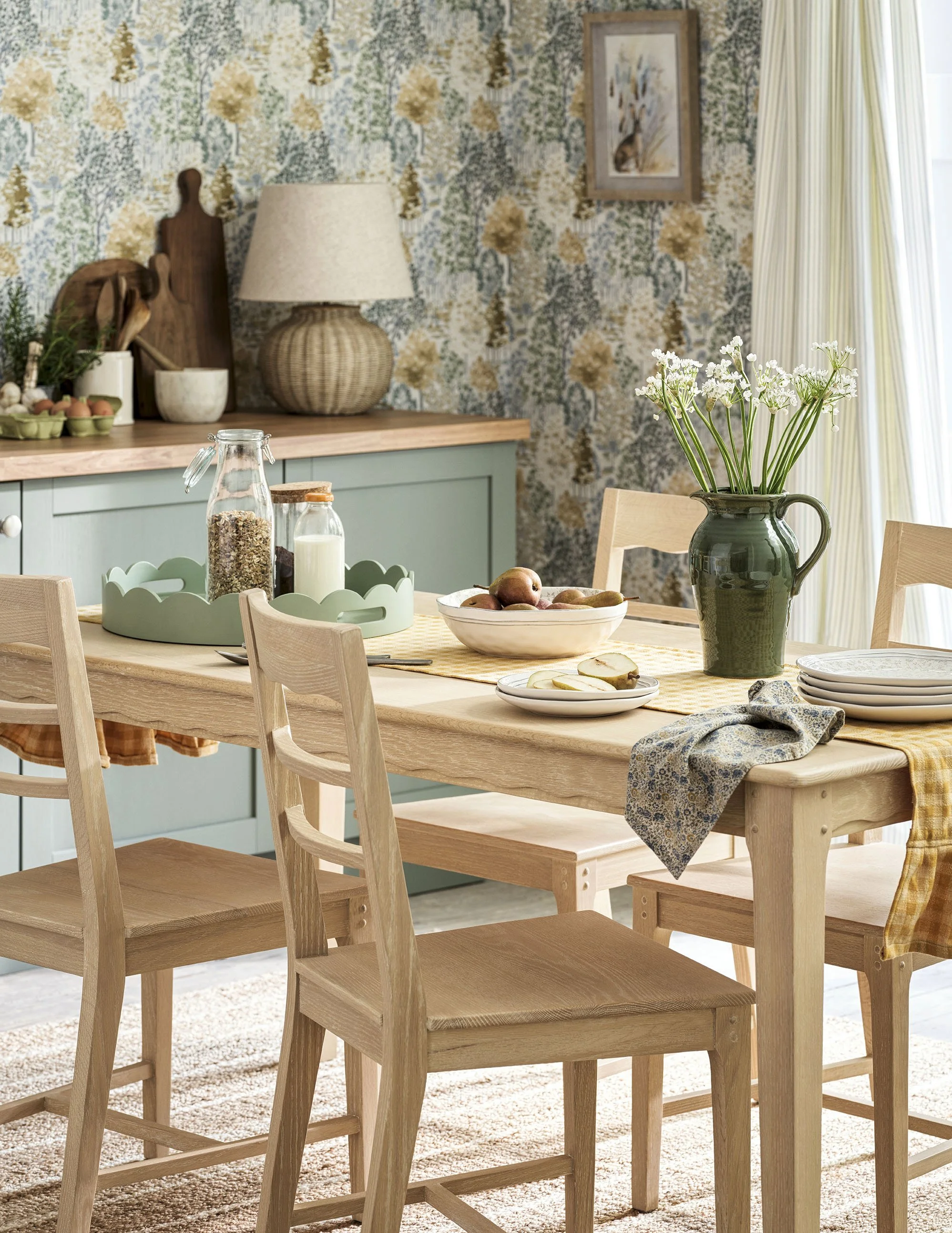 A cozy dining room with a wooden table set with plates, a bowl of apples, and a green vase with white flowers. Light wood chairs surround the table, and a sideboard with a lamp, decorative items, and framed artwork is in the background, all set again