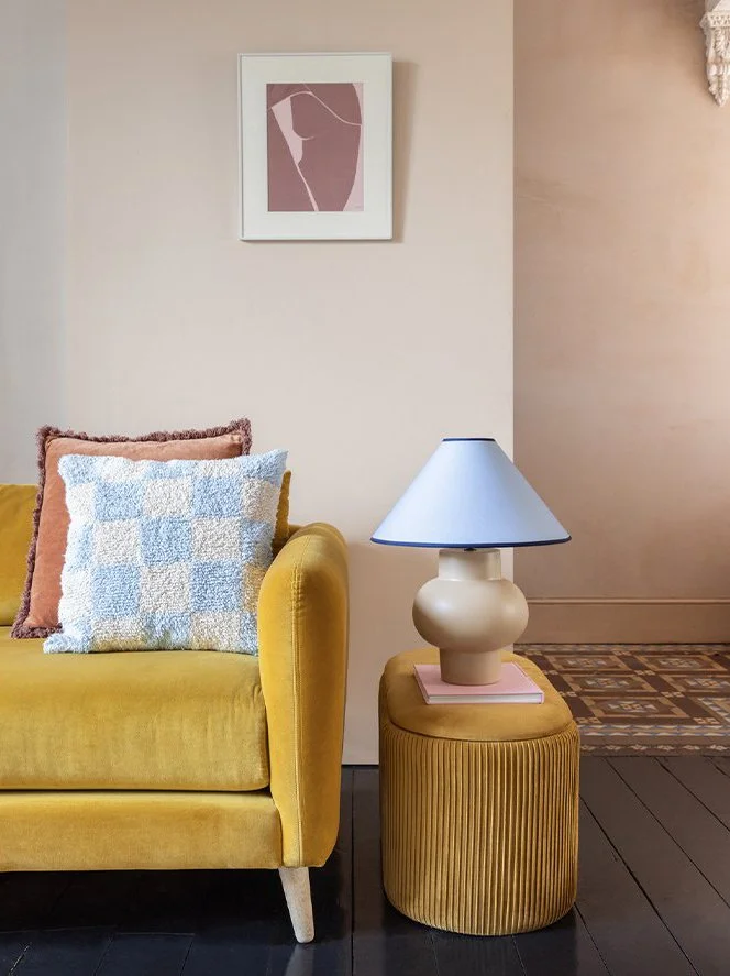 Close-up of a yellow velvet sofa with two decorative pillows, one pink and one blue with a textured pattern, next to a yellow ottoman holding a beige lamp with a blue shade, set against a beige wall with minimalist artwork.
