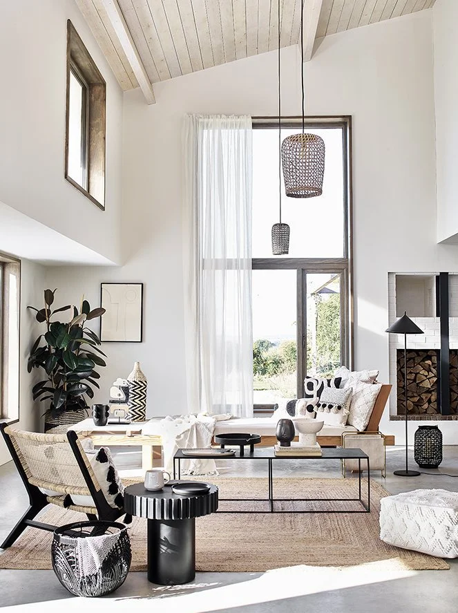 Modern living room with high vaulted ceiling, large window with sheer curtains, and Scandinavian-inspired decor. Features a beige sofa, black and white accents, a wood fireplace, and various decorative objects on tables and shelves.