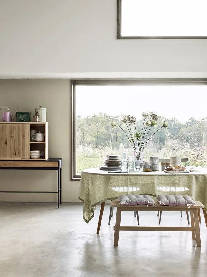 A cozy dining area with a round table covered with a light green tablecloth, set with plates, glasses, and a loaf of bread. Two cushioned benches are positioned at each side of the table. Large windows in the background reveal an outdoor natural land