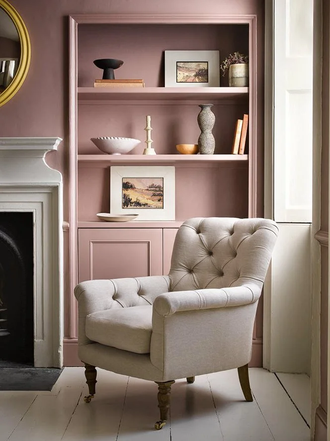 A beige tufted armchair with wooden legs sits in front of a pink built-in bookshelf, which contains decorative items and framed artwork. A white fireplace is on the left, and a window with sunlight is on the right.