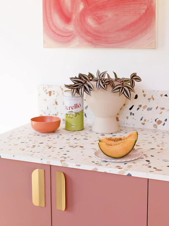 A kitchen counter with a pink cabinet, a white terrazzo countertop, a white vase with a plant, a melon slice on a small plate, a canned Perello artichoke, a pink bowl, and a white mug with a floral pattern. A pink abstract painting hangs on the white