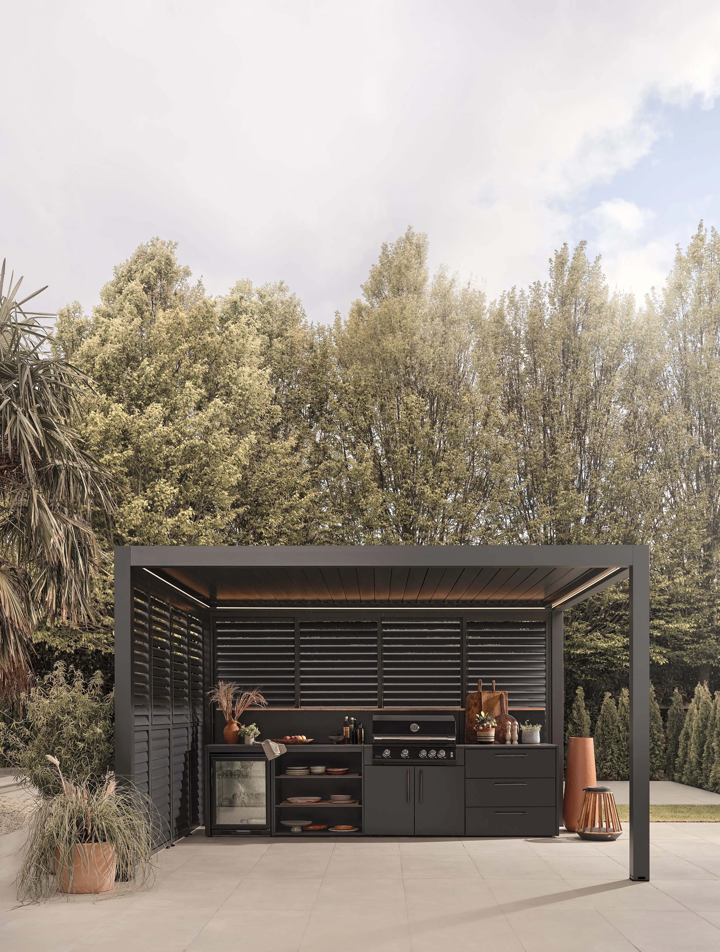 Outdoor kitchen with black cabinets, a grill, and decorative plants, set on a patio with trees in the background.