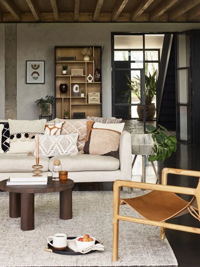 Modern living room with a beige sofa decorated with patterned pillows, a round wooden coffee table with decorative items, a black bookshelf with plants and art, large glass doors leading to a plant-filled outdoor area, and a leather chair with a wood