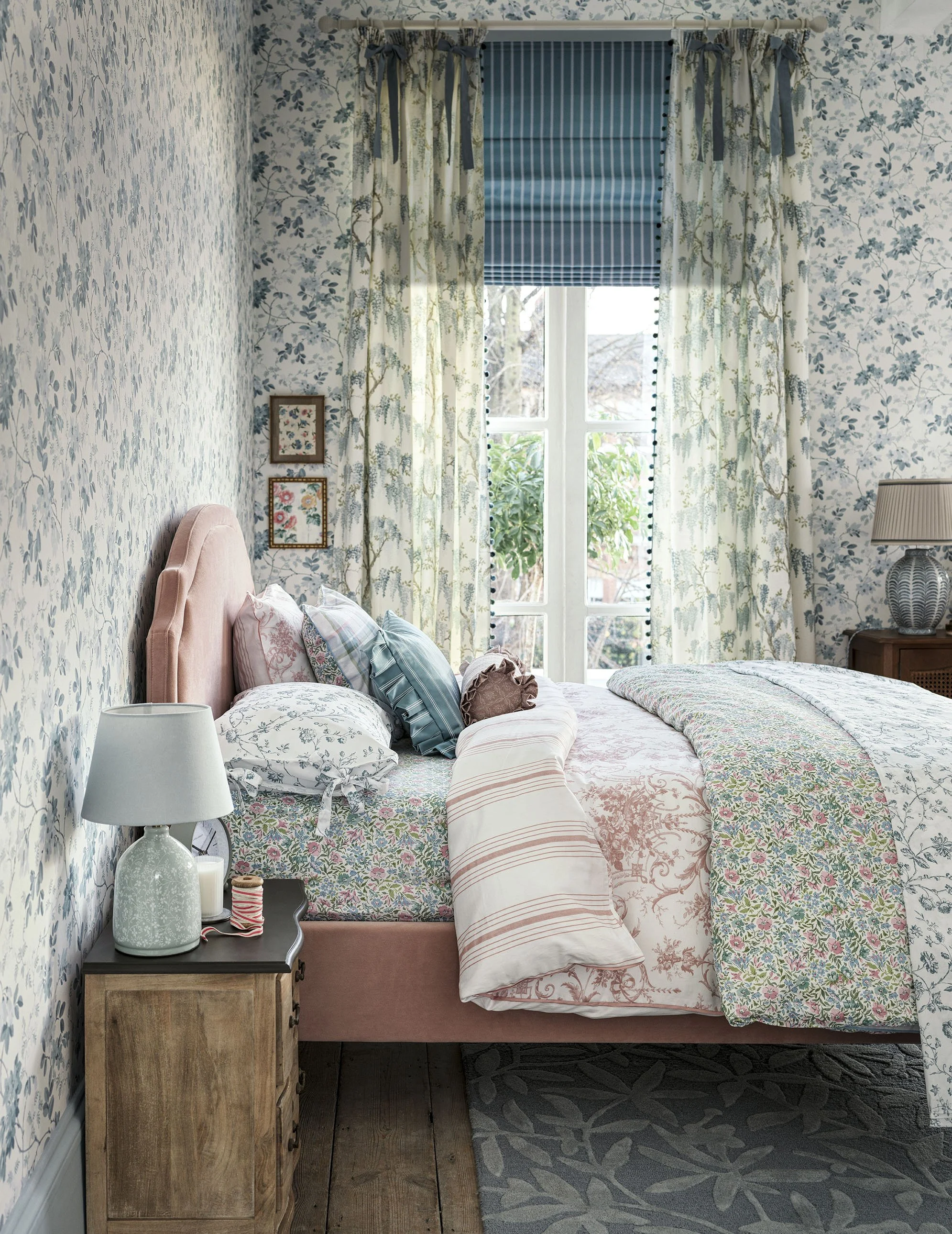 A cozy bedroom with floral wallpaper, a pink upholstered bed, multiple pillows, a nightstand with a lamp, and a window with curtains and blinds showing outdoor greenery.