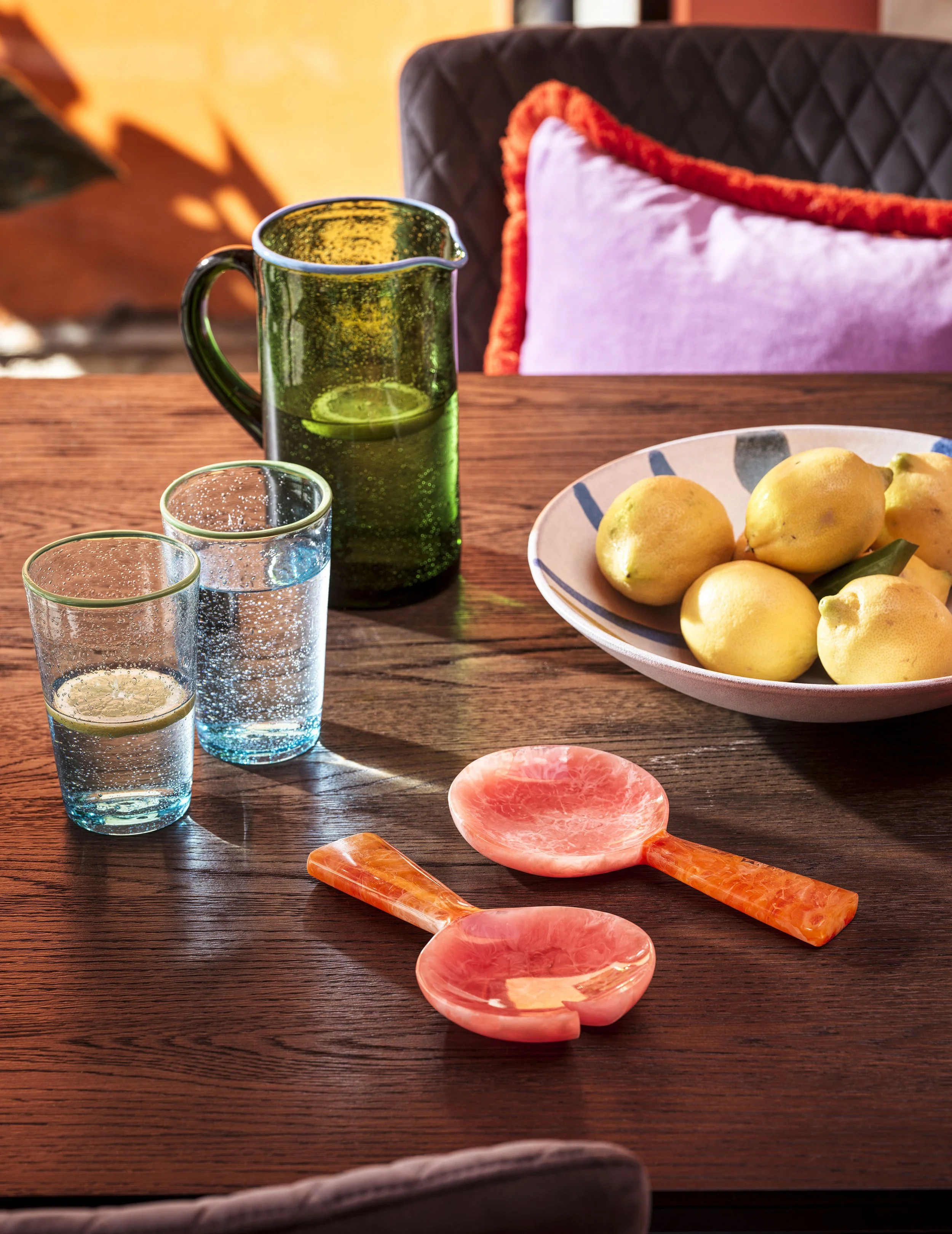 A wooden dining table with a green glass pitcher of water with lemon slices, two glasses of sparkling water with lemon, a white bowl of lemons, and a pink marble spoon and spatula, with a dark cushioned chair and pink pillow in the background.