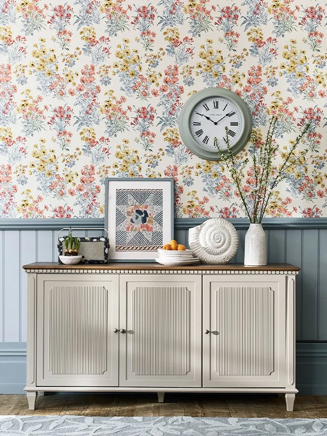 A traditional sideboard against a floral wallpapered wall with a large clock. The sideboard has decorative items including framed artwork, a plant in a pot, a bowl of oranges, a large shell sculpture, and a tall vase with branches.