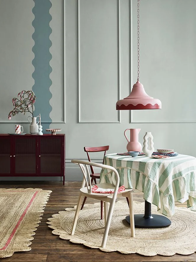 A dining room with a table covered by a green and white checkered tablecloth, decorated with various vases, bowls, and plates. There are two chairs, one red and one white, positioned around the table. A pink hanging lamp illuminates the space. In the