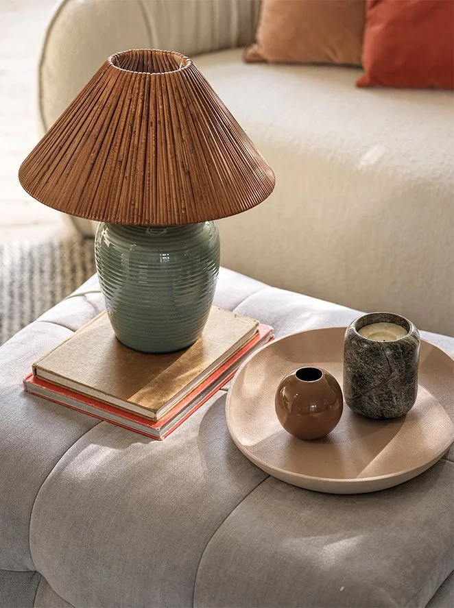 A living room table with a ceramic lamp, stacked notebooks, and a tray holding two small vases, with a beige couch and pillows in the background.
