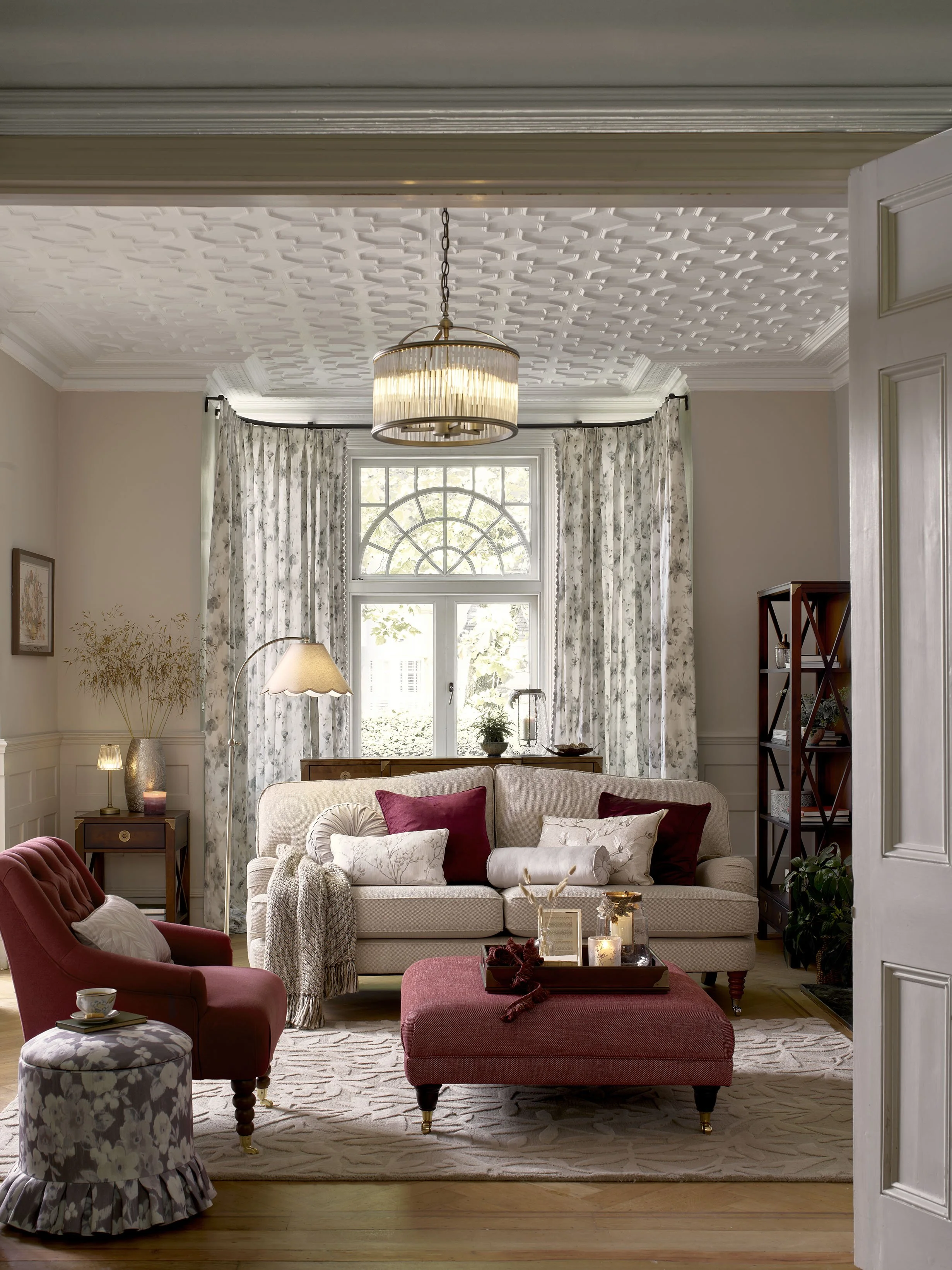 A cozy living room with a beige sofa, red armchair, ottoman, and patterned curtains in front of large windows, decorated with pillows, candles, and indoor plants.