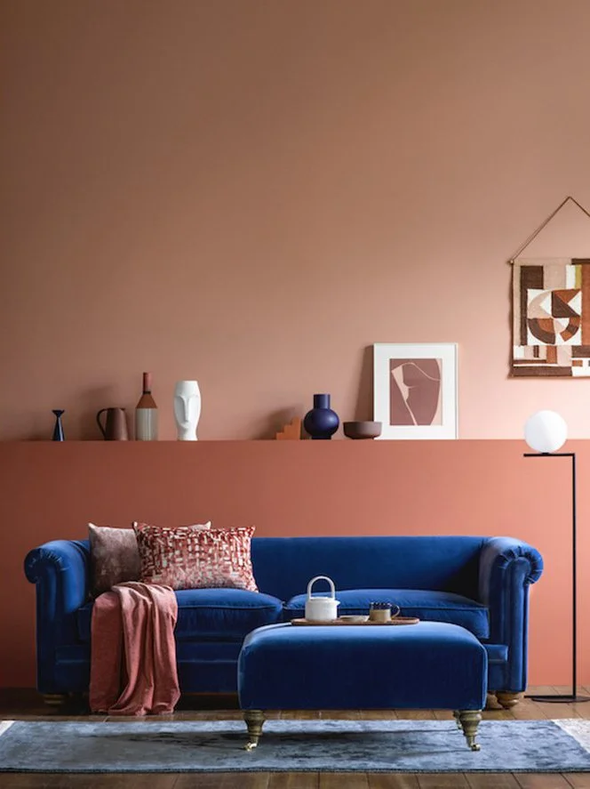 Living room with a blue velvet sofa, pinkish wall, various decorative vases on a ledge, framed artwork, woven wall hanging, a white lamp on a black stand, and a tray with a teapot on the sofa.