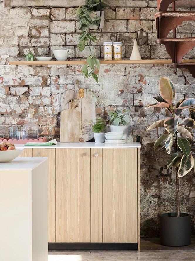 Kitchen corner with exposed brick wall, wooden shelf with jars and dishes, light wooden cabinet, countertop with bowls and a cutting board, potted plants, and a decorative staircase.